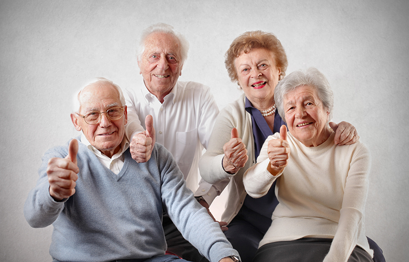 Four spirited seniors celebrate friendship with joyful smiles and thumbs up, embodying a life filled with laughter and shared memories behind a soft backdrop.